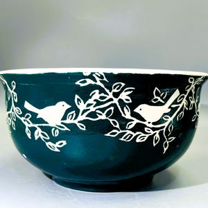 May include: A teal ceramic bowl with a white rim and a white bird and branch design. The bowl has a rounded shape and is suitable for serving or display. The bird and branch design is repeated around the bowl.