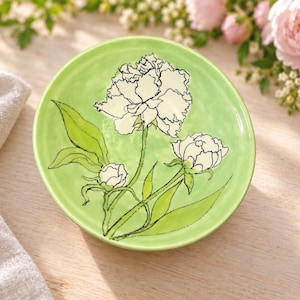 May include: A round, light green ceramic dish featuring a hand-drawn illustration of white peony flowers with black outlines. The surface of the dish has a subtle, wavy texture, and the floral design includes two blooming flowers and one bud.