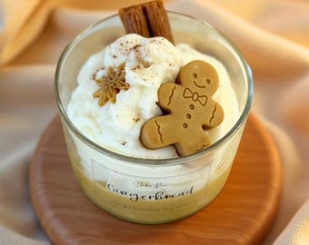 Puede incluir: Una vela de vidrio con temática de pan de jengibre, con una galleta de hombre de jengibre, una rama de canela y anís estrellado sobre una cera batida. La vela está sobre un posavasos de madera, con la palabra "Gingerbread" en la etiqueta.