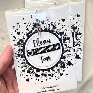 Könnte beinhalten: Ein weißer runder Schlüsselanhänger mit einem schwarz-weißen Musiknoten-Design. Der Schlüsselanhänger verfügt über einen schwarz-weißen Barcode mit den Namen "Elena" und "Tom", die über und unter dem Barcode geschrieben sind.