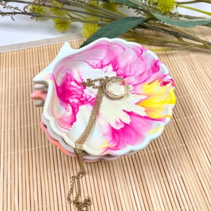 Könnte beinhalten: Ein Stapel muschelförmiger Schmuckschalen mit einem lebendigen rosa, gelben und weißen Blumendesign. Eine goldene Halskette und ein Ring liegen auf der obersten Schale. Die Schalen sind auf einer Bambusmatte gestapelt.
