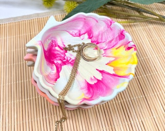Könnte beinhalten: Ein Stapel muschelförmiger Schmuckschalen mit einem lebendigen rosa, gelben und weißen Blumendesign. Eine goldene Halskette und ein Ring liegen auf der obersten Schale. Die Schalen sind auf einer Bambusmatte gestapelt.