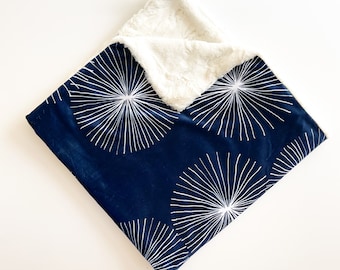 Pode incluir: Um cobertor azul marinho para bebé com um padrão branco de explosão estelar de um lado e um material de pelúcia macia creme do outro. O cobertor é quadrado e dobrado.
