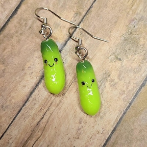 May include: A pair of dangle earrings featuring green and yellow pickle-shaped charms with a smiling face. The earrings have silver hooks.