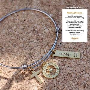 Puede incluir: Una pulsera de brazalete plateada con dijes dorados. Los dijes incluyen una cruz, un colgante circular y una etiqueta rectangular que dice "even if". La pulsera se muestra sobre una superficie texturizada de color marrón claro.