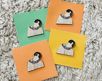 May include: Four enamel pins featuring a person with black hair and a white sweater with a blue and red collar. Each pin is placed on a different colored square: green, orange, and yellow. The background is a fluffy, gray surface.