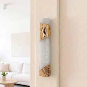 May include: A rectangular mezuzah case featuring a combination of translucent white resin and natural wood grain sections. A gold-colored metal Hebrew letter Shin is centered on the upper wooden portion of the vertical, elongated object.