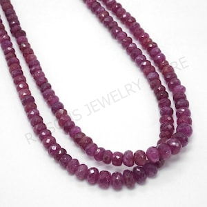 May include: Two strands of faceted ruby beads in varying shades of red and pink. The beads are strung together to form a necklace. The gemstones have a polished, reflective surface. The background is white.