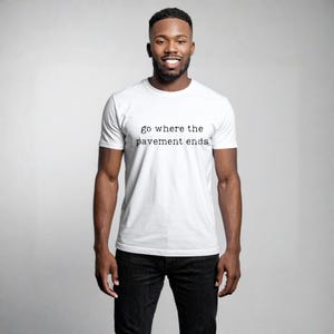 Puede incluir: Una camiseta blanca con la frase "go where the pavement ends" impresa en negro. La camiseta es de manga corta y la lleva una persona. La persona lleva pantalones negros.