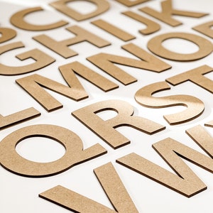 May include: A set of wooden letters in a light brown color, arranged in a random order on a white surface. The letters are all uppercase and include the letters A through Z.