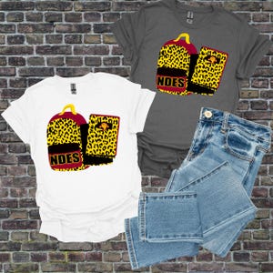 May include: White and grey t-shirts with a graphic of a backpack and notebook featuring a yellow leopard print design. The backpack and notebook have the text "NDES" in red. Light blue denim jeans are also in the image.