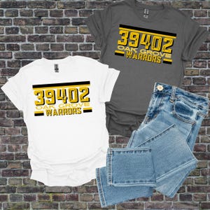 Puede incluir: Dos camisetas, una blanca y otra gris, con el texto "39402 OAK GROVE WARRIORS" en amarillo y negro. Un par de vaqueros azules está doblado junto a las camisas. Las camisetas son probablemente para una escuela o un equipo.