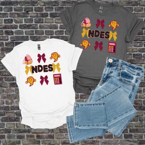 May include: Two t-shirts, one white and one grey, with a school-themed design. The design includes a backpack, bows, a tornado, a calculator, and the word "NDES". A pair of blue jeans is also in the image.