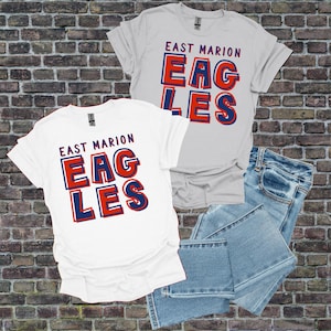 May include: Two white t-shirts and a pair of blue jeans are displayed against a brick wall. The shirts feature the text "EAST MARION EAGLES" in red and blue block letters. One shirt is light gray.