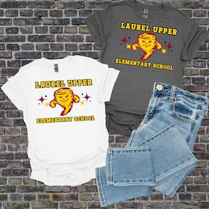 Op de afbeelding: Twee t-shirts, een witte en een grijze, met de tekst "LAUREL UPPER ELEMENTARY SCHOOL" en een cartoon tornado-afbeelding. Een spijkerbroek staat ook op de afbeelding.