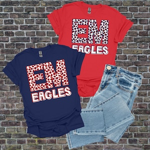 May include: Two short-sleeved t-shirts, one red and one navy blue, with the text "EM EAGLES". The letters "EM" are filled with a red and white leopard print. Light blue denim jeans are folded next to the shirts.