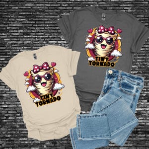 May include: Two t-shirts, one beige and one gray, featuring a cartoon tornado design with sunglasses, a bow, and the words "Tiny Tornado." Light blue denim jeans are folded next to the gray shirt.