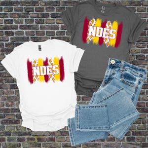 Peut inclure: Deux t-shirts, un blanc et un gris, avec le mot "NDES" en gras, accentué de coups de pinceau rouges et jaunes. Une paire de jeans bleus est également dans l'image. Les t-shirts ont un col rond et des manches courtes.