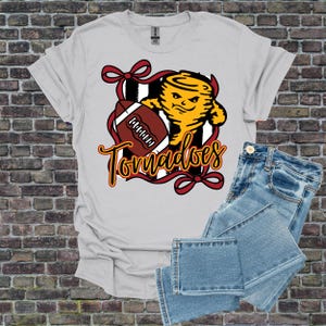 Pode incluir: Uma camiseta cinza claro com um gráfico de um tornado amarelo segurando uma bola de futebol, com a palavra "Tornadoes" em uma fonte cursiva. O design é emoldurado por uma fita vermelha. Jeans de ganga azul claro dobrados ao lado da camiseta.