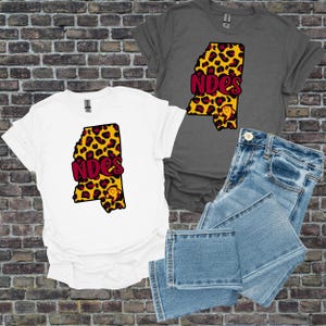 May include: Two t-shirts, one white and one grey, featuring a leopard print Mississippi state outline with the text "NDES" in maroon. A pair of blue jeans is also visible. The shirts have a graphic of a monkey.