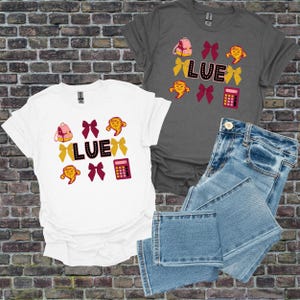 Op de afbeelding: Witte en grijze t-shirts met het woord "LUE" in zwarte letters, versierd met een roze rugzak, gele strikken, een rekenmachine en een stripfiguur. Een spijkerbroek staat ook op de foto.
