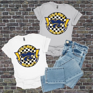 May include: Two t-shirts, one white and one grey, displaying a Columbia Wildcats logo with a blue wildcat, yellow lightning bolts, and a chequered background. Light blue denim jeans are folded beside the shirts. The shirts are on a brick background.