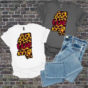 Op de afbeelding: Witte en grijze t-shirts met een luipaardprint omtrek van de staat Mississippi. Het woord "Love" is gedrukt in bordeauxrood, en een cartoon leeuw staat onderaan. Een blauwe jeans is ook in de afbeelding.