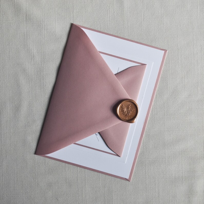 Puede incluir: Un sobre rosa polvoriento sellado con un sello de cera dorado con un dise&ntilde;o de hoja, sobre una tarjeta blanca con un borde rosa a juego. El sobre est&aacute; parcialmente abierto, revelando la tarjeta en su interior.