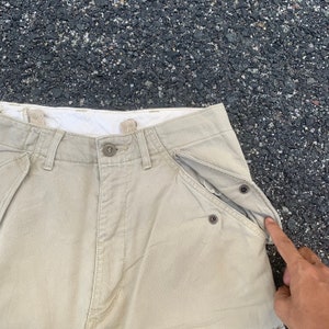 May include: Khaki-colored pants with button and zipper closure. The pants have multiple pockets, including a unique pocket design with snap closures. The fabric appears to be a durable, woven material.