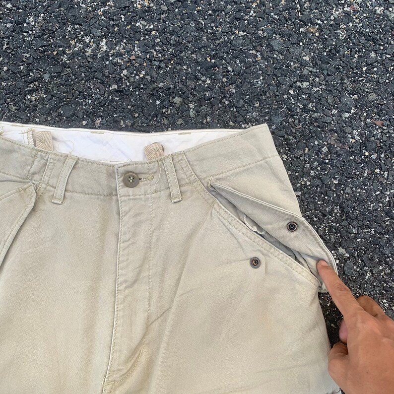 May include: Khaki-colored pants with button and zipper closure. The pants have multiple pockets, including a unique pocket design with snap closures. The fabric appears to be a durable, woven material.