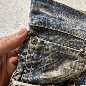 May include: Close-up of a section of blue denim jeans. The jeans show signs of wear with faded areas and distressed details. Visible are a pocket, metal rivets, and stitching. The fabric has a textured appearance.