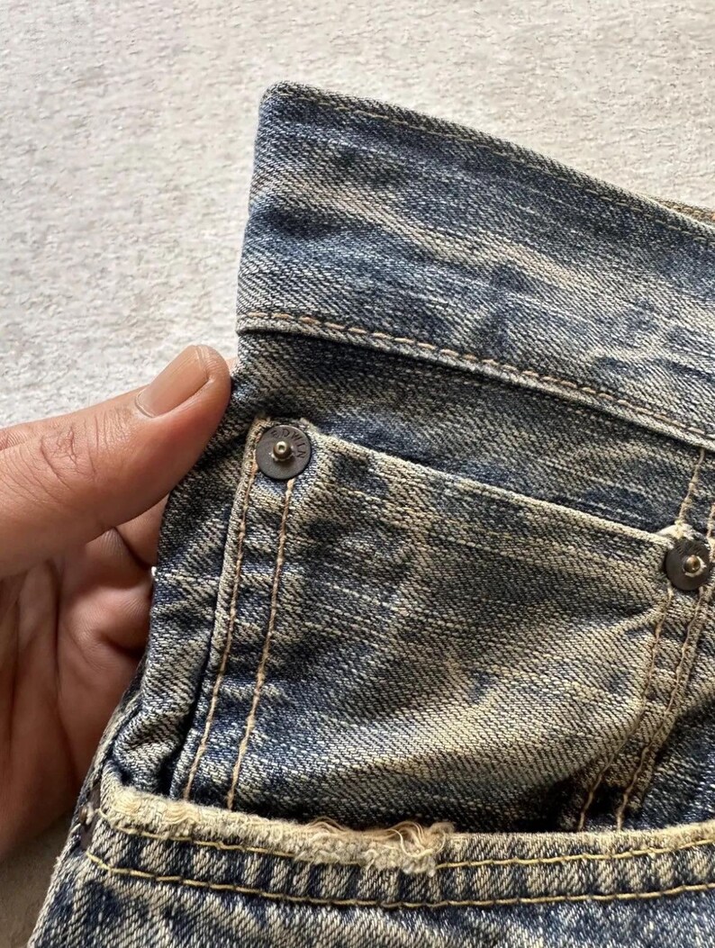 May include: Close-up of a section of blue denim jeans. The jeans show signs of wear with faded areas and distressed details. Visible are a pocket, metal rivets, and stitching. The fabric has a textured appearance.