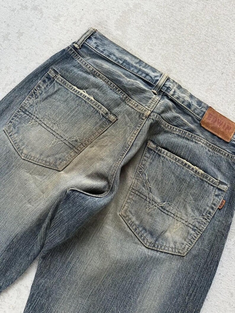 May include: Close-up of the back of faded blue denim jeans. The jeans have two back pockets with decorative stitching and a brown leather patch with the brand name "EDWIN". The denim has a worn, distressed look.