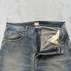 May include: Faded blue denim jeans with a button and zip fastening. The jeans have a classic five-pocket design and show signs of wear, with a slightly distressed appearance. The fabric has a textured look.