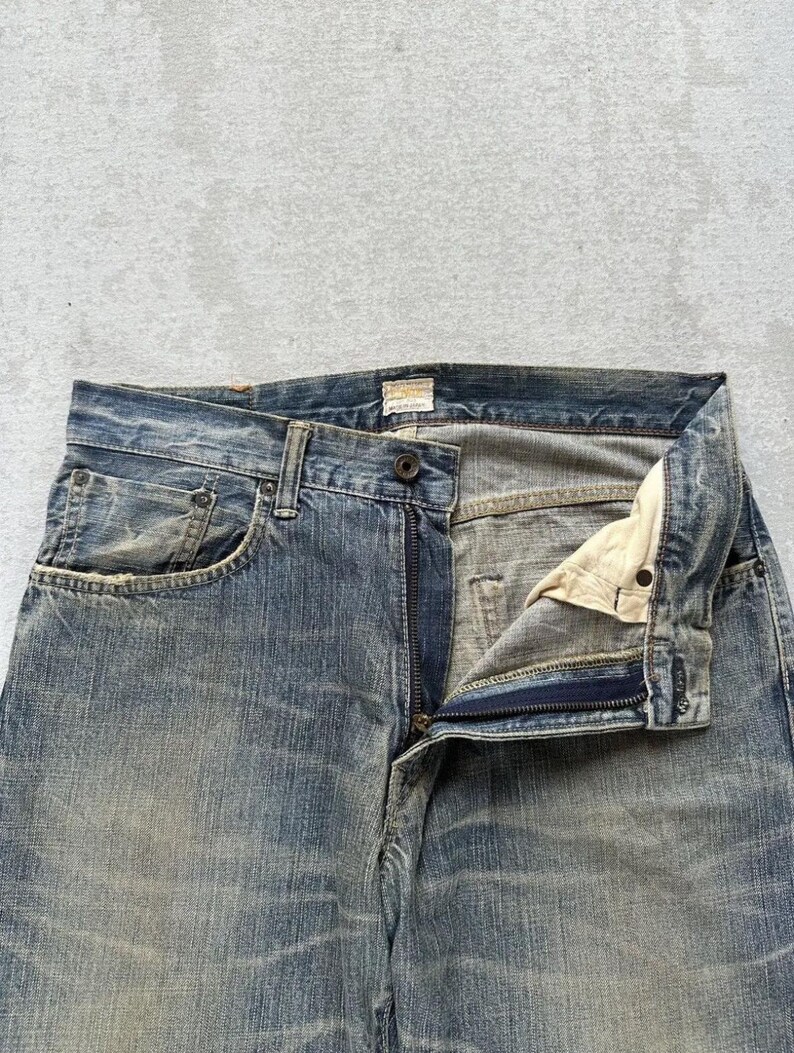 May include: Faded blue denim jeans with a button and zip fastening. The jeans have a classic five-pocket design and show signs of wear, with a slightly distressed appearance. The fabric has a textured look.