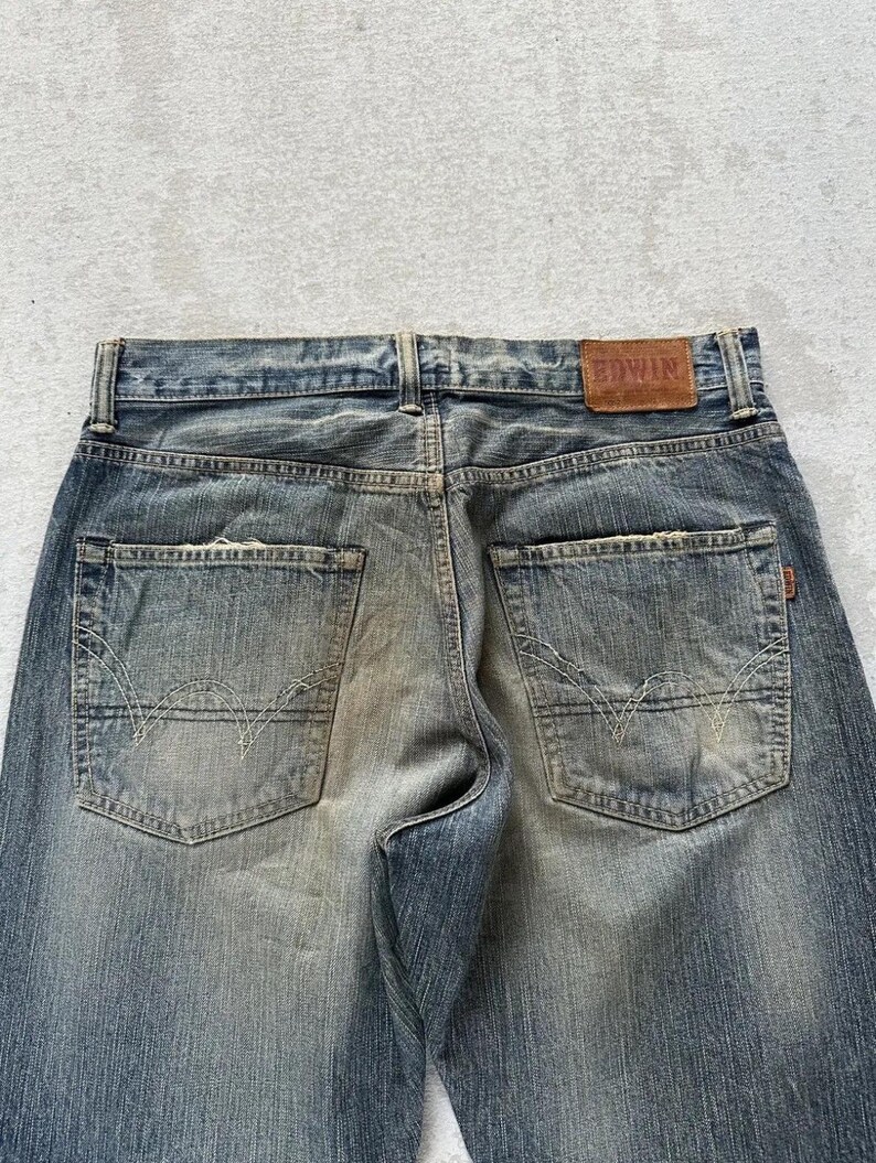 May include: Faded blue denim jeans with a classic five-pocket design. The back pockets feature a distinctive stitch pattern. A brown leather patch with the brand name "EDWIN" is visible at the waistband.