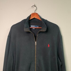 May include: Black zip-up jacket with a stand-up collar and a small red logo on the chest. The jacket has a full zipper closure and is hanging on a wooden hanger. The Polo Ralph Lauren logo is visible on the inside of the collar.