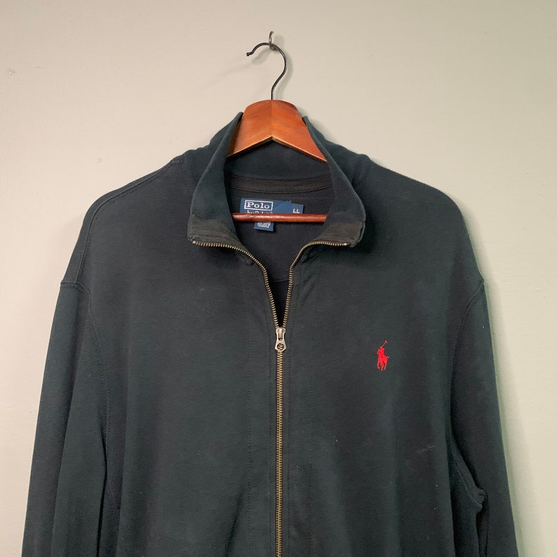 May include: Black zip-up jacket with a stand-up collar and a small red logo on the chest. The jacket has a full zipper closure and is hanging on a wooden hanger. The Polo Ralph Lauren logo is visible on the inside of the collar.