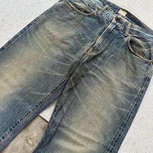 May include: Faded blue denim jeans with a classic five-pocket design. The jeans show signs of wear and tear, with lighter patches and subtle distressing throughout the fabric. The jeans have a button and zip closure.