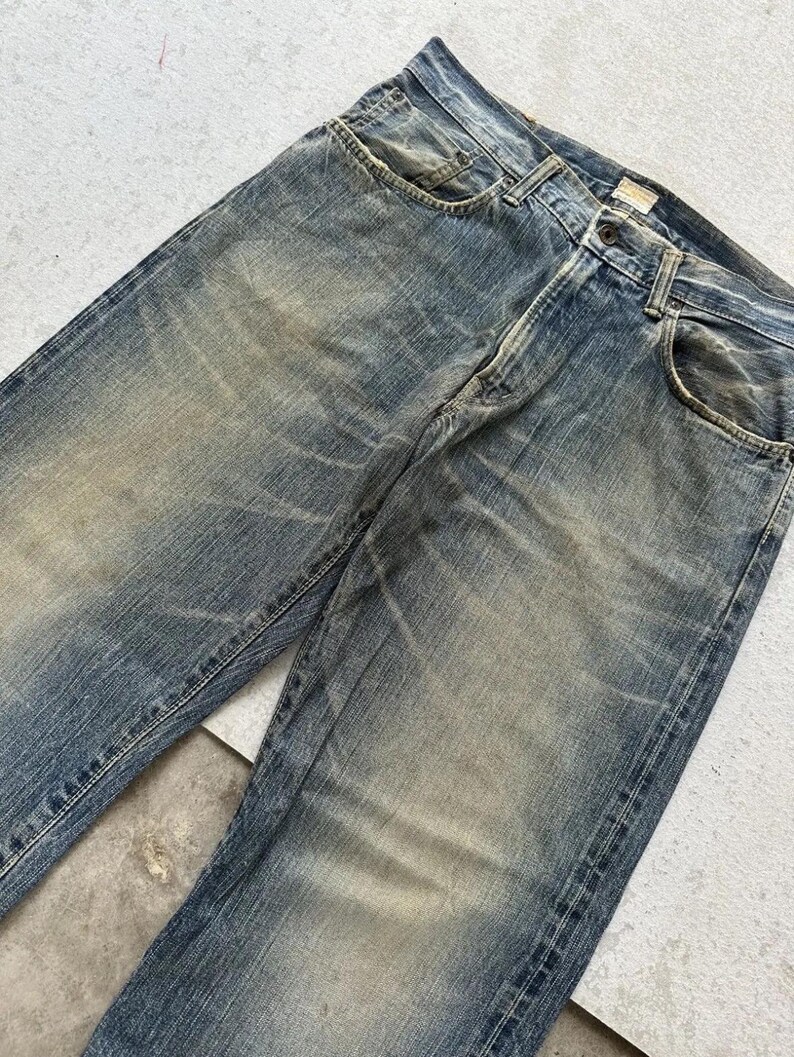 May include: Faded blue denim jeans with a classic five-pocket design. The jeans show signs of wear and tear, with lighter patches and subtle distressing throughout the fabric. The jeans have a button and zip closure.