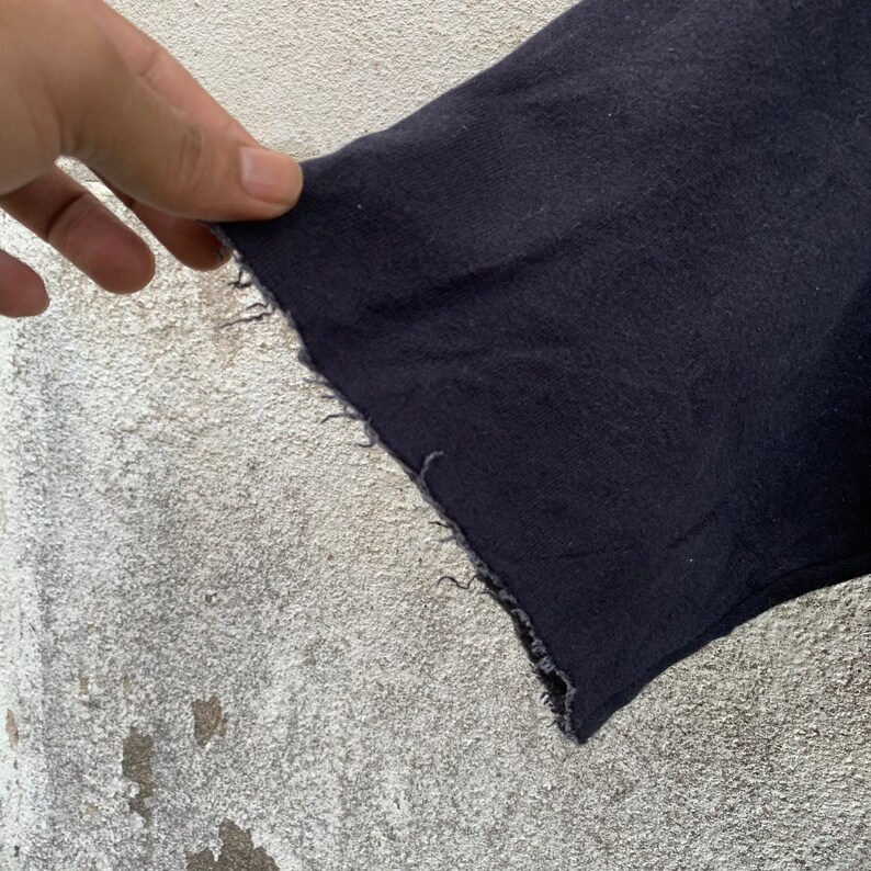 May include: Close-up of a dark navy blue fabric with a raw, unfinished edge. The fabric appears to be a section of a garment, possibly a sleeve or hem, with visible frayed threads along the cut edge.