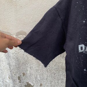 May include: Close-up of a dark navy blue t-shirt sleeve with a distressed, raw-edge hem. The shirt features a celestial design with the letter "D" visible in silver. The fabric appears worn and vintage.