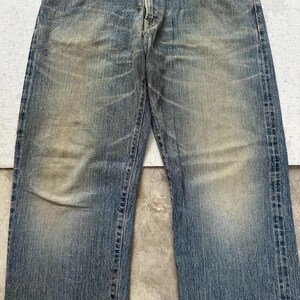 May include: Faded blue denim jeans with a classic five-pocket design. The jeans show signs of wear with lighter patches and subtle distressing throughout the fabric. The straight-leg cut offers a timeless style.