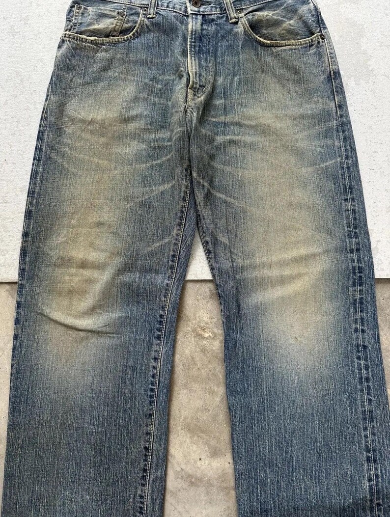 May include: Faded blue denim jeans with a classic five-pocket design. The jeans show signs of wear with lighter patches and subtle distressing throughout the fabric. The straight-leg cut offers a timeless style.
