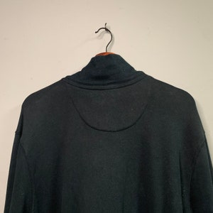 May include: A black sweatshirt with a high collar, hanging on a wooden hanger. The back of the sweatshirt is visible, showing a stitched design near the collar. The fabric appears to be a soft, comfortable material.