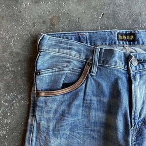 May include: Close-up of blue denim jeans with a faded wash. The jeans feature a brown leather trim around the pocket and a "BLUE TRIP" label. The fabric shows subtle wear and tear.