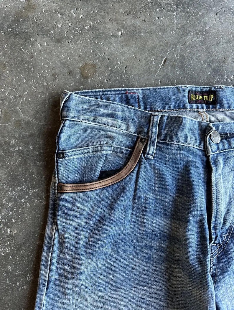May include: Close-up of blue denim jeans with a faded wash. The jeans feature a brown leather trim around the pocket and a "BLUE TRIP" label. The fabric shows subtle wear and tear.