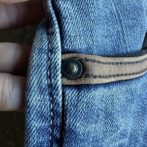 May include: Close-up of blue denim fabric with a brown leather accent and a metal button. The denim has a textured appearance, and the button is riveted to the fabric. The leather accent is stitched to the denim.