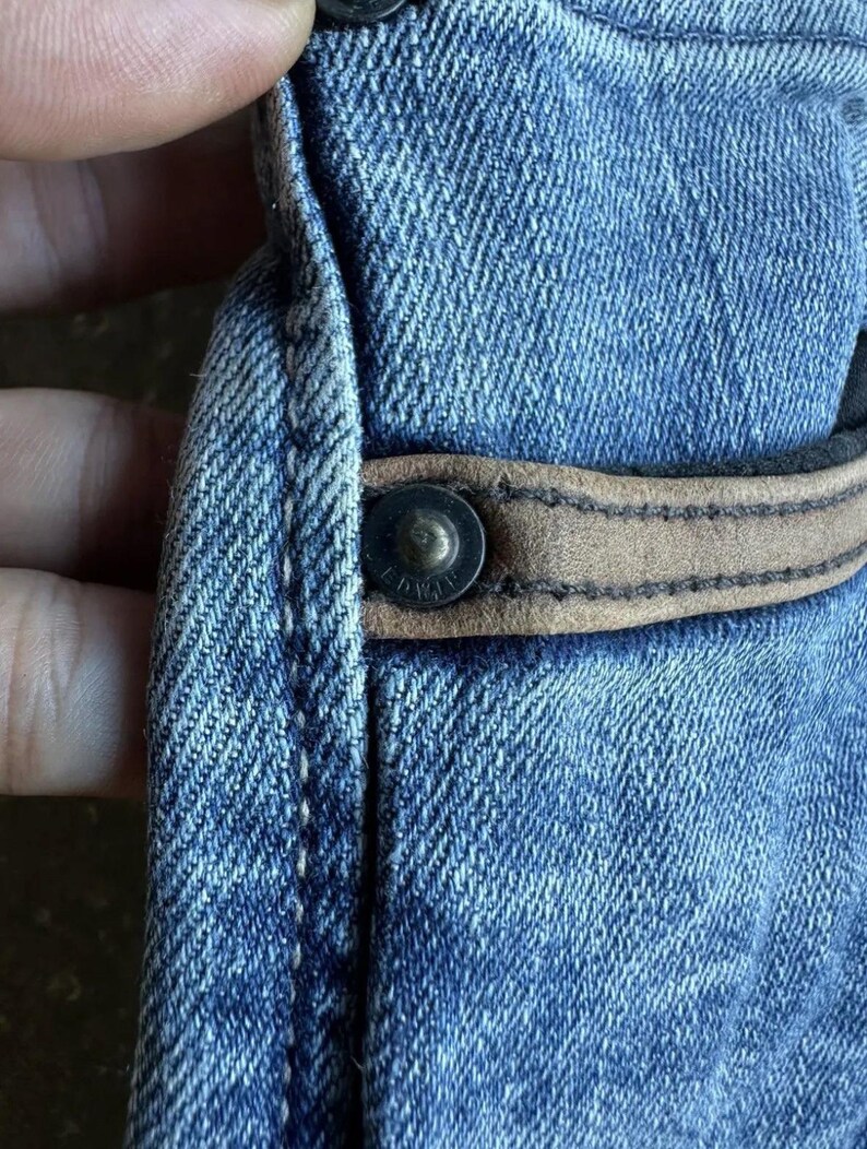 May include: Close-up of blue denim fabric with a brown leather accent and a metal button. The denim has a textured appearance, and the button is riveted to the fabric. The leather accent is stitched to the denim.