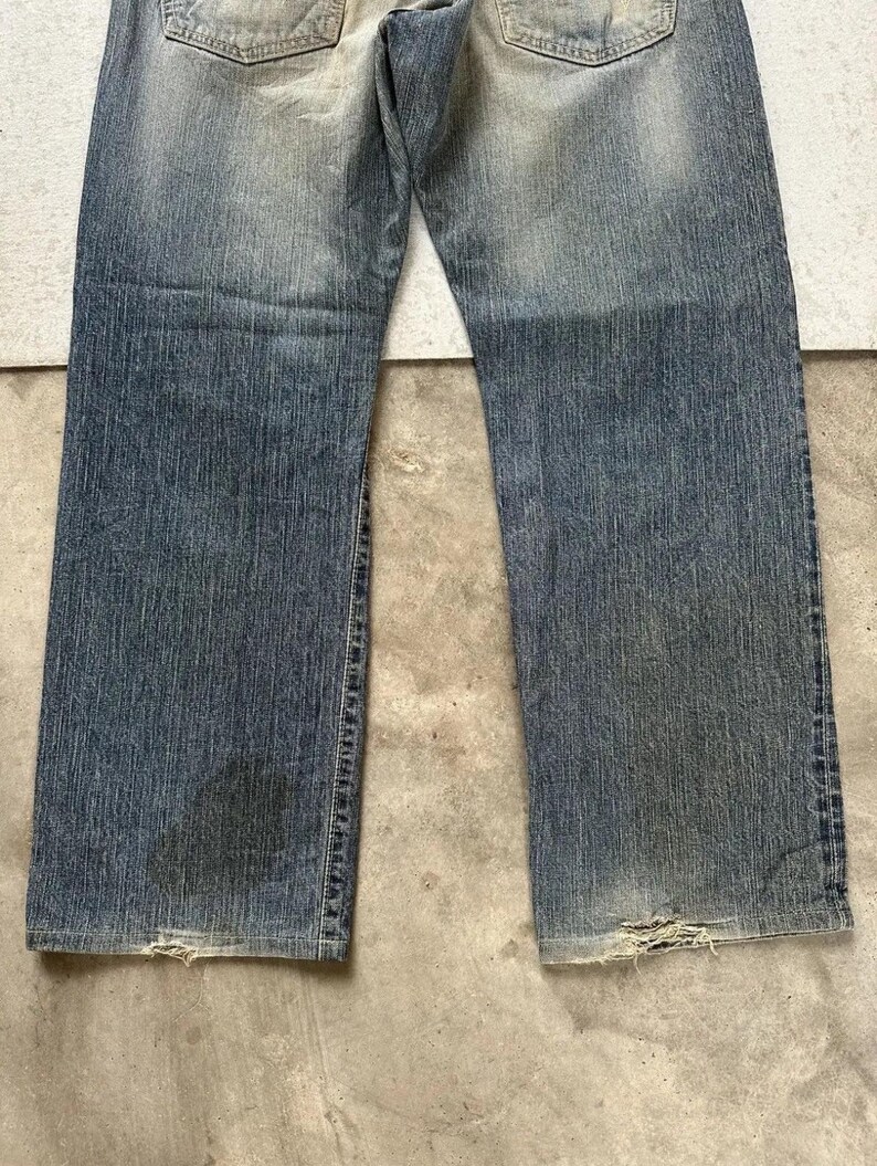 May include: Faded blue denim jeans with a worn appearance. The jeans show signs of wear and tear, with distressed areas near the bottom hems. The denim has a gradient effect, with lighter shades at the top and darker shades towards the bottom.